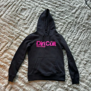 Svart hoodie från Dirt Cult - Säljer en svart hoodie från Dirt Cult med rosa text 'Dirt Cult San Diego' på bröstet. Den har en klassisk känguruficka och en mysig huva. Perfekt för en avslappnad stil eller en kylig dag. 🖤💗