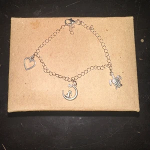 Silverfärgat armband med berlocker - Säljer ett superfint silverfärgat armband med tre olika berlocker: ett hjärta, en måne med en stjärna och en liten sköldpadda. Perfekt för att ge en personlig touch till din stil. Armbandet har en justerbar kedja och stängs med en karbinhake.