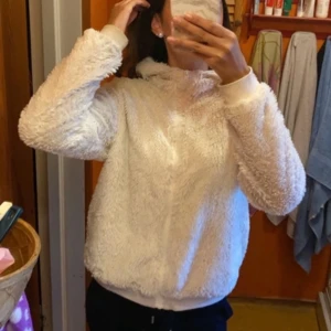 Vit teddyjacka från KappAhl - Mysig och fluffig vit teddyjacka från KappAhl. Perfekt för kyliga dagar med sin mjuka och varma känsla. Jackan har en dragkedja framtill och en huva för extra komfort. Den är långärmad och passar perfekt för både höst och vinter. Super fint skick 