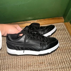 Svarta sneakers från Puma - Säljer ett par snygga svarta sneakers från Puma i storlek 39. Skorna är i skinn med en vit sula och har klassisk snörning. Perfekta för både vardag och träning. De är i nytt skick och redo för en ny ägare!