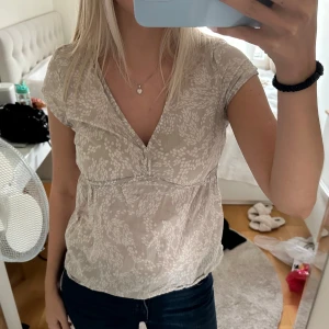 Beige blommig blus - Säljer en söt beige/grå blus med blommigt mönster. Den har en v-ringad design och korta ärmar, perfekt för en avslappnad look. Blusen är i ett lätt och luftigt material, vilket gör den bekväm att bära under varma dagar.