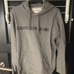 Grå hoodie från Calvin Klein Jeans - Säljer en stilren grå hoodie från Calvin Klein Jeans med logga på bröstet. Tröjan har en klassisk design med huva och dragsko samt en praktisk magficka. Perfekt för en avslappnad look.