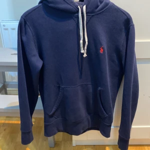Mörkblå hoodie från Ralph Lauren - Säljer en snygg mörkblå hoodie från Ralph Lauren med klassisk logga på bröstet. Tröjan har en justerbar huva med vita snören och en praktisk magficka. Perfekt för en avslappnad stil.