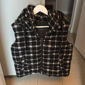 Svartvit rutig väst från Zara - Snygg svartvit rutig väst från Zara med dragkedja och huva. Perfekt för lager-på-lager under kyligare dagar. Västen har en modern och stilren design som passar till många olika outfits.