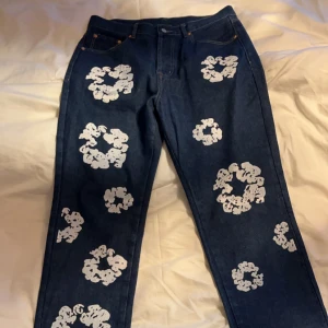 Levi's 501 jeans Denim Tears collab - Unika Levi's 501 jeans i blå denim med ett coolt vitt blommönster över hela byxorna. Klassisk femficksdesign med knappgylf och raka ben. Perfekt för dig som gillar en lite mer baggy / relaxed fit.
