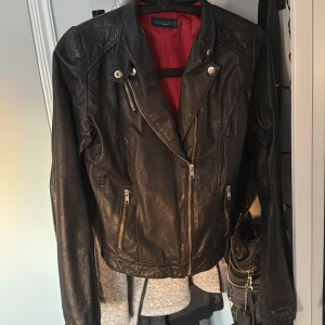 Svart skinnjacka - Snygg svart skinnjacka med coola dragkedjedetaljer och en röd insida. Köpt på Sellpy. Hittar ingen storlek men skulle säga att den är typ en S