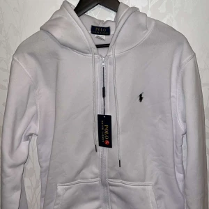 Äkta Vit hoodie från Polo Ralph Lauren - Snygg vit hoodie från Polo Ralph Lauren med dragkedja och klassisk logga på bröstet. Perfekt för en stilren och avslappnad look. Den har en justerbar huva och praktiska fickor framtill.