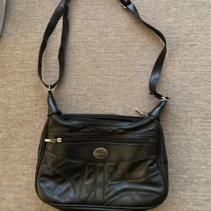 Svart axelväska i skinn - Snygg svart axelväska i skinn med justerbar axelrem och flera dragkedjefickor. Perfekt för att hålla ordning på dina saker med stil.