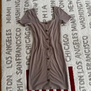 Beige ribbad klänning från Divided - Snygg beige klänning från Divided med ribbad struktur och knappar längs hela framsidan. Den har korta ärmar och en figurnära passform som framhäver silhuetten. Perfekt för en stilren look.