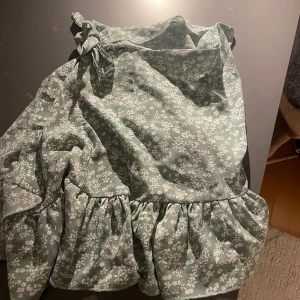 Grön blommig klänning - Charmig grön klänning med blommigt mönster och volangdetaljer. Den har en knytning vid axeln och en luftig passform, perfekt för varmare dagar.