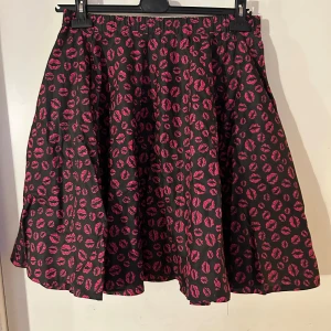 Svart kjol med rosa läppmönster - Trendig svart kortkjol med ett iögonfallande rosa läppmönster. Perfekt för att ge din outfit en lekfull touch. Kjolen har en bekväm passform och är lätt att matcha med olika toppar. Helklockad. Resår i midjan