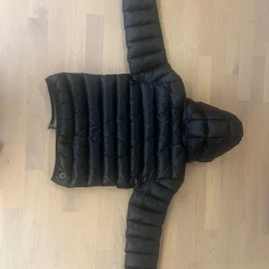 Svart dunjacka från Colmar Stlk 12 , 154 cm  - Säljer en svart dunjacka från Colmar med huva. Jackan har en quiltad design och är perfekt för kyligare väder. Den har långa ärmar och två fickor med dragkedja framtill. Storlek 12 YR,  154 cm. Hel, ren och inga revor. 