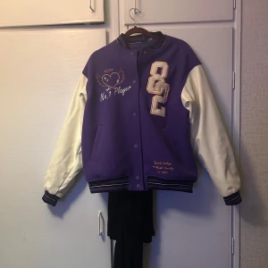 Lila och vit bomberjacka - Snygg lila bomberjacka med vita ärmar och broderade detaljer. Jackan har knappar framtill och ribbade muddar vid ärmslut och nederkant. Perfekt för en cool och avslappnad stil.