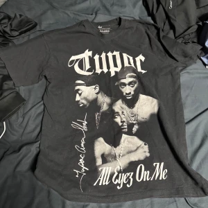 Svart T-shirt med Tupac-tryck från Pull&Bear - Säljer en svart T-shirt från Pull&Bear med ett stort tryck av Tupac och texten 'All Eyez On Me'. Perfekt för fans av hiphop och streetwear. T-shirten är kortärmad och gjord i bomull.
