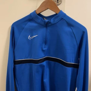 Blå långärmad tröja från Nike - Storlek 158-170. Snygg blå långärmad tröja från Nike med dragkedja och vit randdetalj över bröstet. Perfekt för träning eller vardagsbruk. Den har en sportig look med en liten Nike-logga på bröstet.