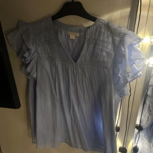 Ljusblå blus med volangdetaljer - Charmig ljusblå blus med volangdetaljer på ärmarna och v-ringad hals. Använd några gånger men inga defekter. Är från neo noir😊