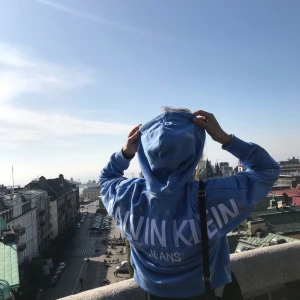 Blå hoodie från Calvin Klein Jeans - Säljer en snygg blå hoodie från Calvin Klein Jeans med stort tryck på ryggen och litet CK-märke fram. Hoodien har en känguruficka och justerbar huva med dragsko. Stor i passformen men relativt kort.