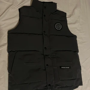 Svart dunväst från Canada Goose - Säljer en stilren svart/grå dunväst från Canada Goose med knappar och broderad logga på bröstet. Västen har en hög krage och praktiska fickor framtill. Perfekt för kyliga dagar.