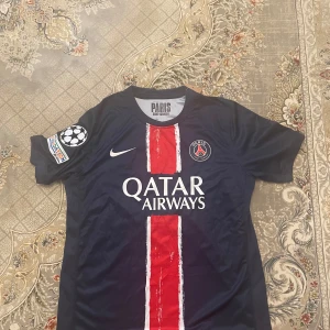 Paris Saint-Germain fotbollströja - Snygg Paris Saint-Germain tröja i mörkblått med röd och vit rand på framsidan. Tröjan har korta ärmar och tryck med 'Qatar Airways' samt klubbens emblem. På baksidan finns namnet 'Mbappé' och nummer 7. Perfekt för fans av PSG!