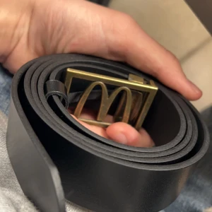 McDonalds bälte 105cm - Snyggt svart bälte med ett unikt guldfärgat spänne. Perfekt för att ge din outfit en stilren touch. Bältet är tillverkat i ett slätt material och har en klassisk design. 105cm långt