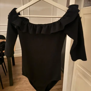 Svart offshoulder topp med volang - Snygg svart offshoulder topp med volangdetaljer runt halsen. Perfekt för en stilren look. Trekvartsärmar 