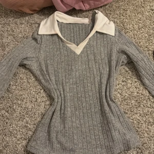 Grå ribbad tröja med vit krage - Snygg grå ribbad tröja med en vit krage och v-ringning. Perfekt för en stilren look. Tröjan har långa ärmar och en figurnära passform.