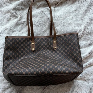 Brun mönstrad axelväska - Snygg brun axelväska med ett klassiskt mönster. Väskan har två långa handtag och en dragkedja upptill. Väldigt rymlig och är perfekt för att bära med sig allt man behöver under dagen🤎