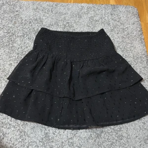 Svart volangkjol med glitterdetaljer - Snygg svart kortkjol med volanger och glittriga detaljer. Perfekt för att ge lite extra bling till din outfit. Den elastiska midjan gör den bekväm och lätt att bära. Lappen med storlek är bortklippt men den är i 164 vilket ungefär motsvarar xs!!