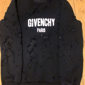 Svart sweatshirt från Givenchy - Snygg svart sweatshirt från Givenchy med vit text 'GIVENCHY PARIS' på bröstet. Tröjan har en unik design med slitna detaljer som ger en edgy look. Perfekt för dig som vill sticka ut med stil.