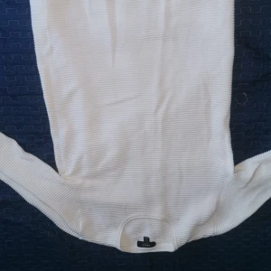 Vit ribbad bodysuit från SMOG - Säljer en stilren vit ribbad bodysuit från SMOG. Perfekt för lager-på-lager eller att bära som den är. Den har en enkel design med knäppning i grenen.