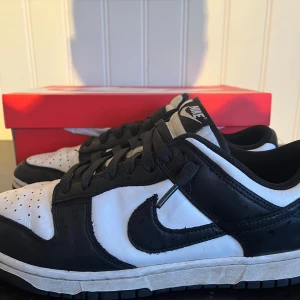Nike Dunk i svart och vitt - Snygga Nike Dunk sneakers i klassisk svart och vit färgkombination. Skorna har en stilren design med snörning och den ikoniska Nike-loggan på sidan. Perfekta för en sportig och trendig look.