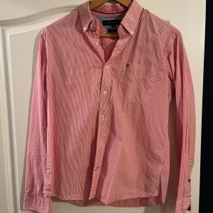 Röd-vit Tommy Hilfiger skjorta. - Säljer en Tommy Hilfiger skjorta som ser rosa ut men den är röd och vit randig. Storleken är XL men jag skulle säga att den passar storlek M. Kom med bud vid intresse, inga returer Tack.