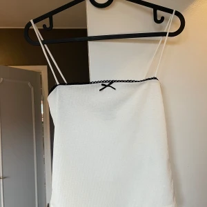 Vit linnetopp från Gina Tricot - Snygg vit linnetopp från Gina Tricot med smala axelband och en söt svart rosett framtill. Perfekt för en stilren och enkel look💗