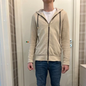 Beige hoodie från Limitato - Säljer en exklusiv beige hoodie från Limitato med ett tryck av Terry O'Neill (James Bond) på ryggen. Hoodien har dragkedja och långa ärmar, perfekt för en stilren look. Begränsad upplaga, nummer 44 av 500. Tröjan är i fint skick köpt på NK för 3795 kr. Tröjan är i storlek Small och modellen på bilden är 170 cm.