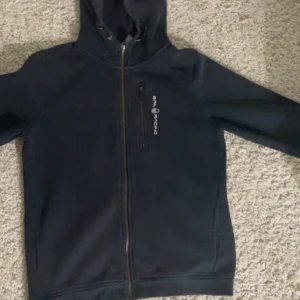 Svart hoodie med dragkedja från Real Racing - Snygg svart hoodie med dragkedja från Real Racing. Den har en praktisk huva och en bröstficka med dragkedja. Perfekt för en avslappnad stil.