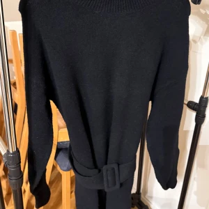 Svart långärmad tröja med bälte - Elegant svart långärmad tröja med ett matchande bälte i midjan. Perfekt för en stilren look. Tröjan har en ribbad halsringning och är tillverkad i ett mjukt material.