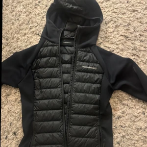 Svart jacka från Peak Performance - Snygg svart jacka från Peak Performance med huva och dragkedja. Jackan har quiltade paneler fram och bak, vilket ger en stilren look. Perfekt för kyligare dagar.