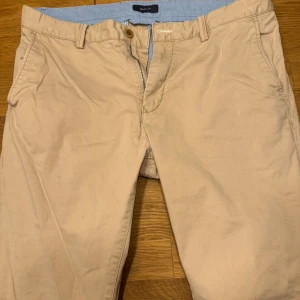 Beige chinos från Gant - Snygga beige chinos från Gant med dragkedja och knappstängning. Klassisk design med två bakfickor och bälteshällor. Perfekta för en stilren look.