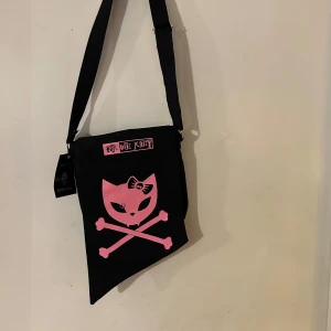 Svart axelväska med rosa kattmotiv - Cool svart axelväska med ett rosa kattmotiv och korsade benknotor. Väskan har en justerbar axelrem och en unik form. Perfekt för att uttrycka en edgy stil.