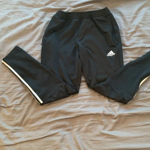 Svarta träningsbyxor från Adidas - Säljer ett par svarta träningsbyxor från Adidas med vita ränder längs sidorna. Byxorna har en elastisk midja och dragkedja vid fickorna. Perfekta för träning eller en avslappnad dag.