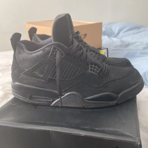 Svarta Nike Air Jordans 4 black cat - Säljer ett par svarta Nike Air Jordans 4 Retro SE. Skorna har en stilren design med snörning och är tillverkade i skinn och syntetmaterial. Perfekta för dig som älskar klassiska sneakers med en modern touch.