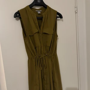 Olivgrön ärmlös klänning - Snygg olivgrön klänning med knytband i midjan för en justerbar passform. Klänningen är ärmlös och har en stilren design som passar perfekt för varma dagar.