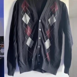 Svart kofta från Dressmann - Snygg svart kofta från Dressmann med klassiskt argyle-mönster i rött och grått på framsidan. Koftan har knappar hela vägen och långa ärmar, perfekt för en stilren look.