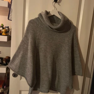 Grå stickad poncho - Mysig grå stickad poncho med polokrage. Perfekt för kyliga dagar och ger en avslappnad look. Den har en lös passform och är enkel att slänga över en tröja eller blus.