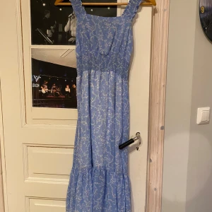 Blå blommig klänning - Charmig blå klänning med blommigt mönster och smockdetaljer i midjan. Klänningen har breda axelband och en luftig passform som ger en romantisk känsla. Perfekt för sommarens varma dagar.