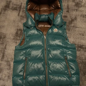 Moncler väst - Snygg grön dunväst från Moncler med dragkedja och avtagbar huva. Västen har två praktiska fickor med dragkedjor och en stilren logga på bröstet. Perfekt för kyligare dagar.storlek 3