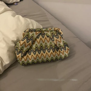 Mönstrad kuvertväska med dragkedja - Säljer en färgglad kuvertväska med zigzag-mönster i beige, blått och grönt. Väskan har en praktisk dragkedja och är perfekt för att bära med sig det viktigaste. Den är lätt att matcha med olika outfits och ger en trendig touch.