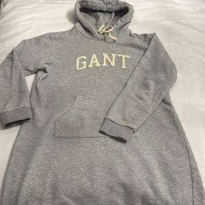 Säljer en stilren grå hoodie-klännimg  från GANT med broderad logga på bröstet. Tröjan har en praktisk magficka och justerbar huva med snören. Perfekt för en avslappnad look.