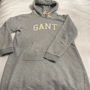 Grå hoodie/klänning från GANT - Säljer en stilren grå hoodie-klännimg  från GANT med broderad logga på bröstet. Tröjan har en praktisk magficka och justerbar huva med snören. Perfekt för en avslappnad look.