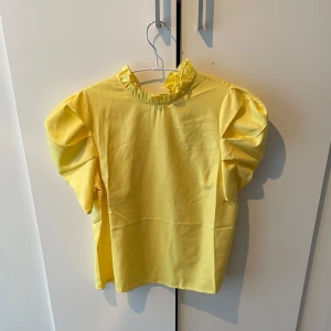 Gul blus med volangdetaljer - Säljer en söt gul blus med volangdetaljer vid halsen och puffiga kortärmar. Perfekt för en färgglad look!